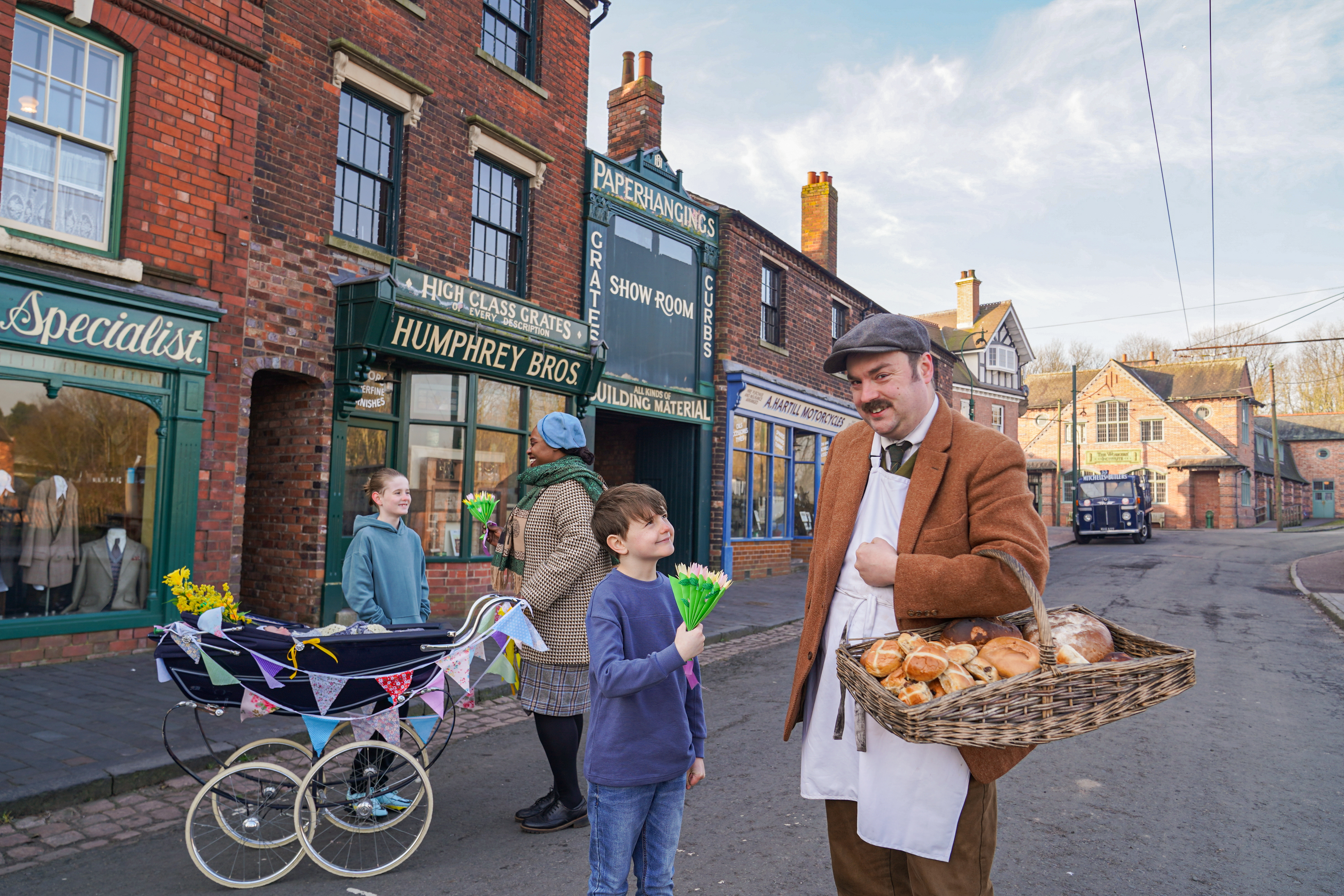 There is a Edwardian Baker stood holding a basket of Hot Cross buns with a young boy of today, behind them stands a 1950s Jamaican mother holding a spring decorated baby pram ready for a carnival parade
