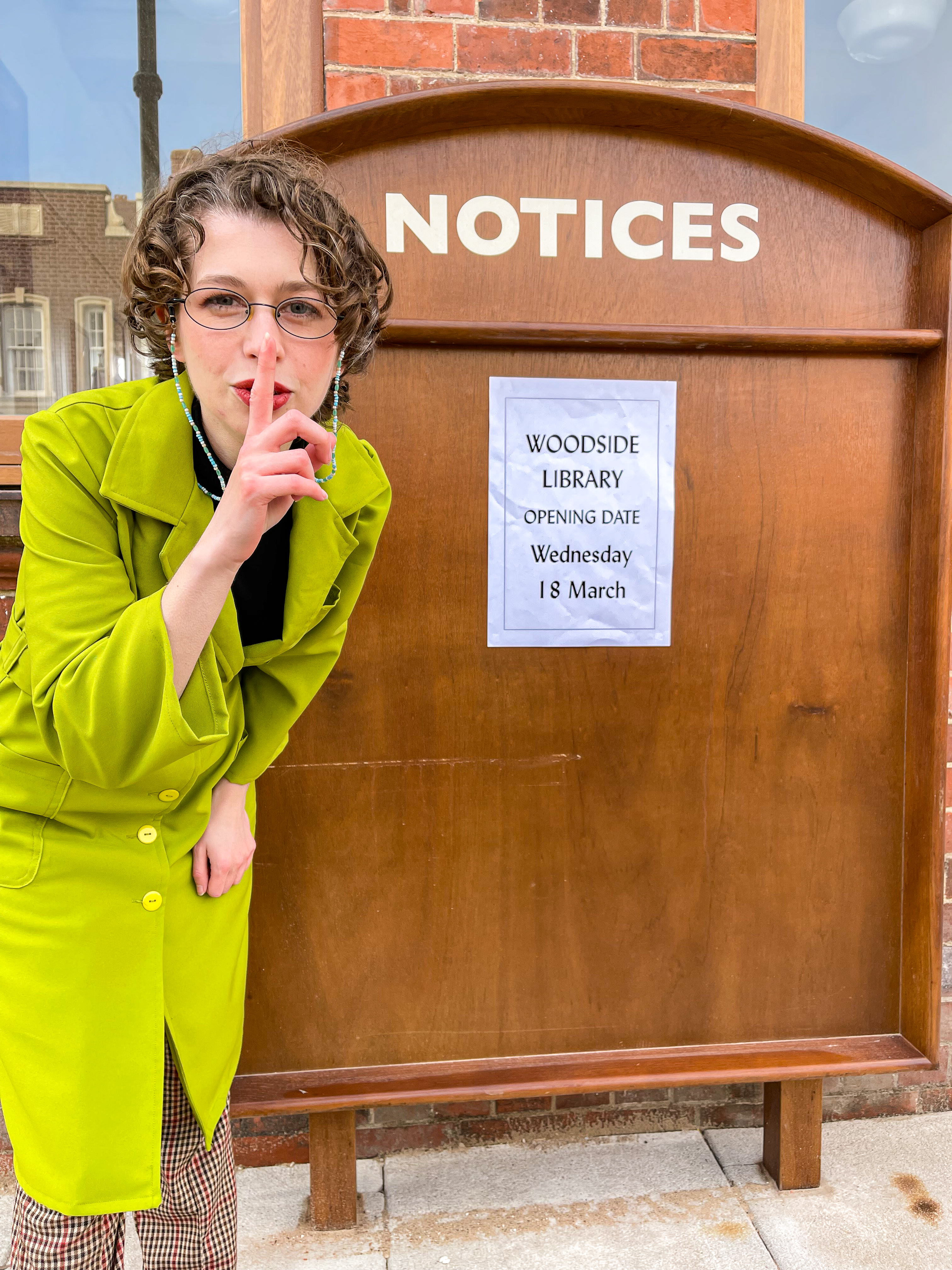 1960s Librarian with her finger to her mouth to say shh, with a notice board sign announcing; Woodside Library opening wednesday 18th March