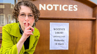 1960s Librarian with her finger to her mouth to say shh, with a notice board sign announcing; Woodside Library opening wednesday 18th March