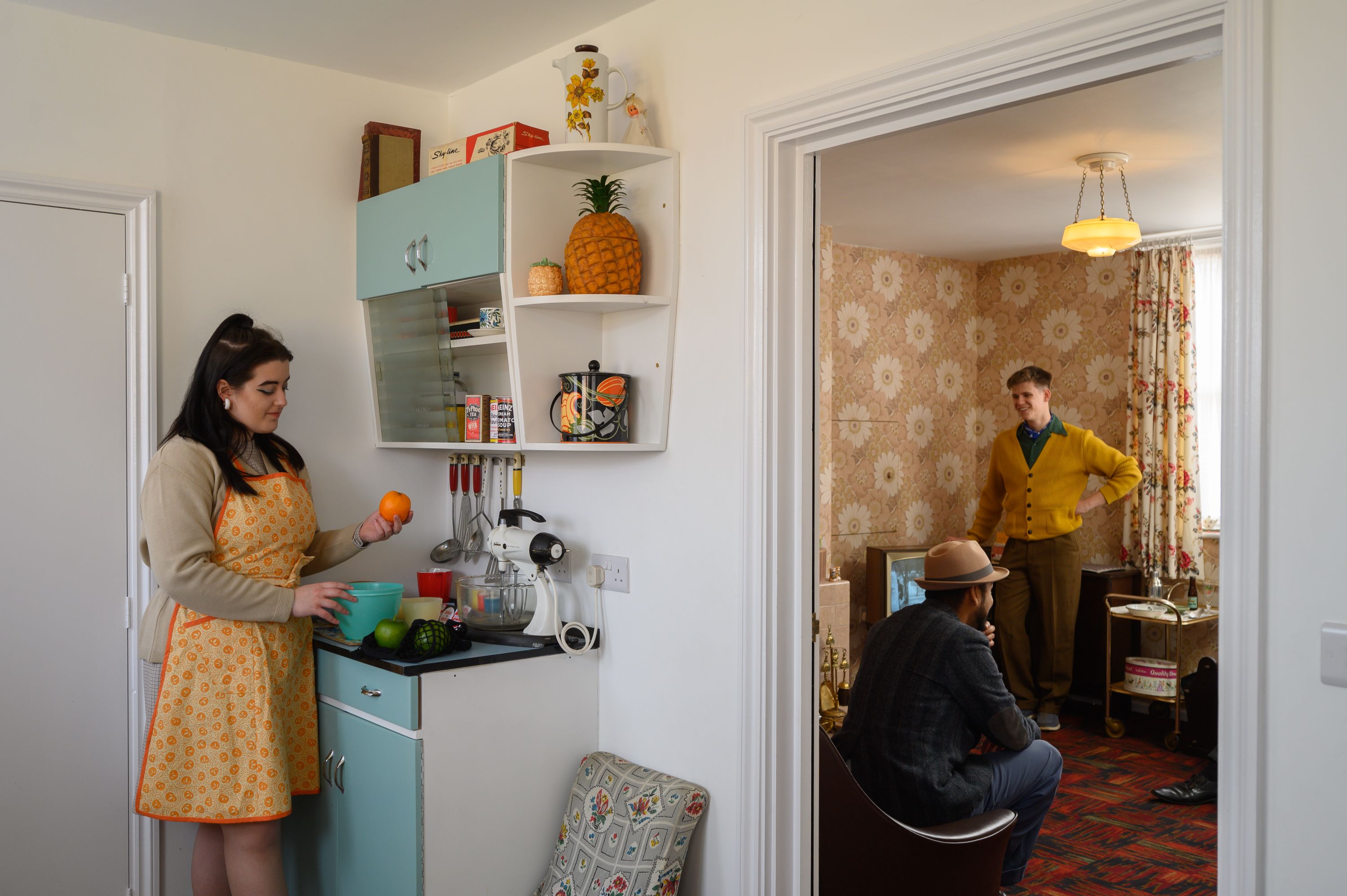 1960s Cast Iron House lady stood in the kitchen, with the door open to the living room where two gents are stood talking. 