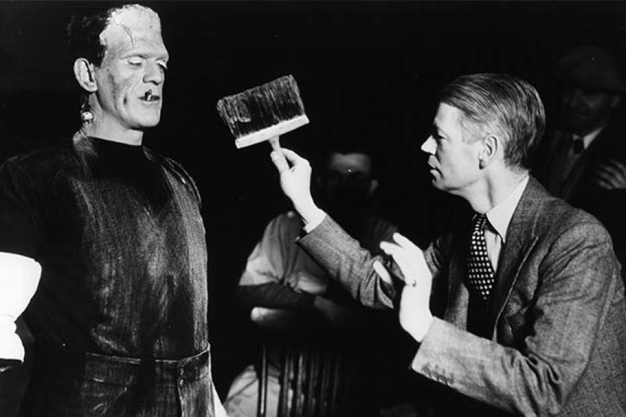 Black and white photograph of Boris Karloff (left) and James Whale (right) on the set of 1931's Frankenstein. Boris is in full costume as Frankenstein, while Whale is talking to him holding a paintbrush.