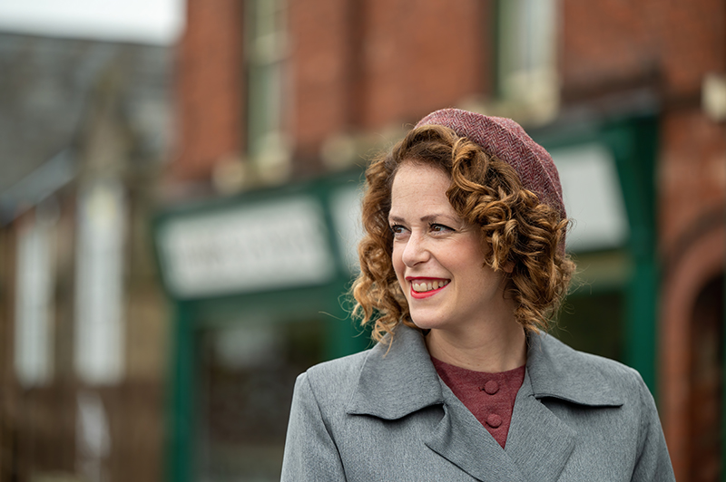 A close up photograph shows a woman dressed in a grey coat wearing a beret that covers her brown curls. She's smiling and stands in front of recreated shops.