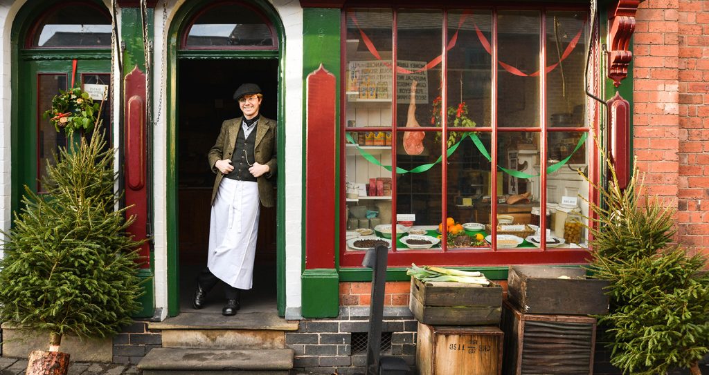 The shop front of Gregory's General Store at Black Country Living Museum. Red and green painted windows display a range of foods and chistmas decorations. A young white man in a white apron and edwardian clothing stands in the doorway. There are Christmas trees and crates of vegetables stacked outside.