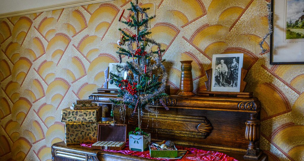 The sideboard in the 1930s apartment. There is an imitation Christmas tree on the side decorated with tinsel and small ornaments. There are family photos and gift wrapped presents next to the tree. The background shows orange and yellow art deco style wallpaper in a fan pattern.