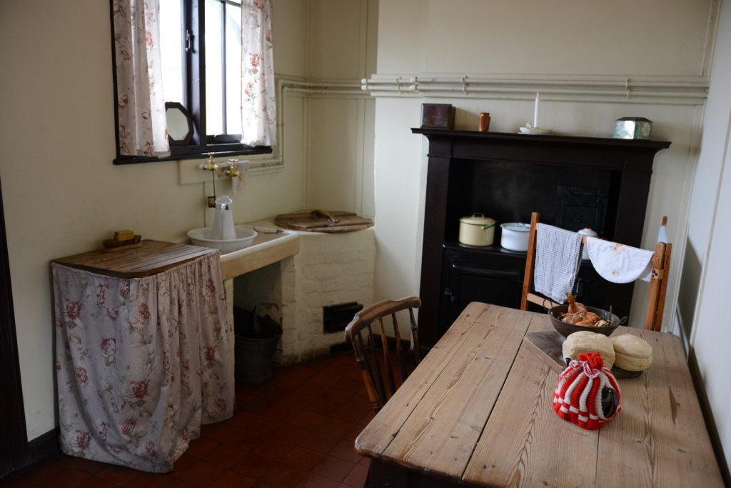 A photograph of the kitchen of the 1940s Cast Iron House. It is quite sparse, with a selection of fixtures and fittings included a small sink and a wooden table with one chair.
